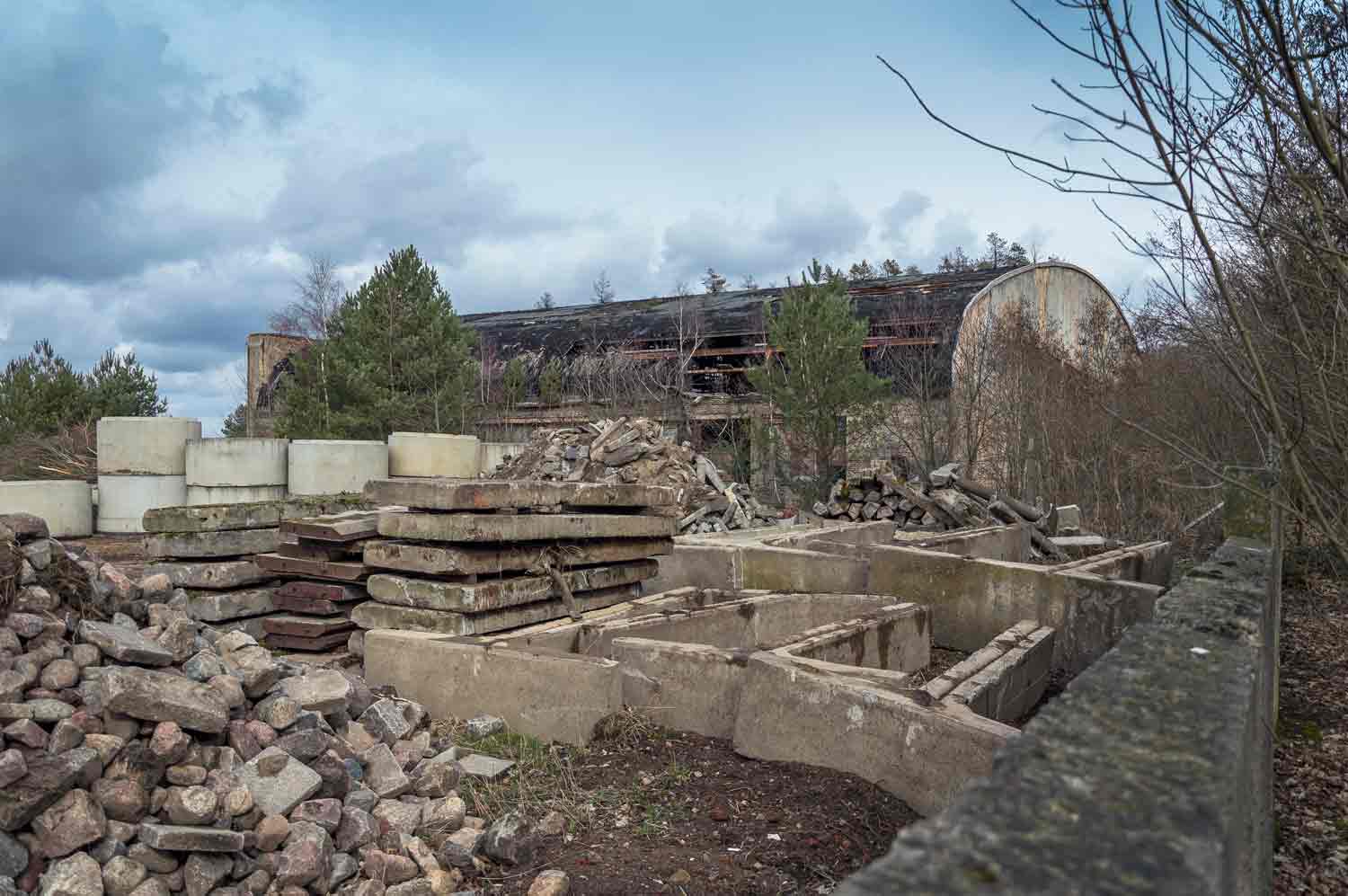 Eingelagerte Betonteile auf einem Gewerbegrundstück, im Hintergrund Ruine eines Hangars
